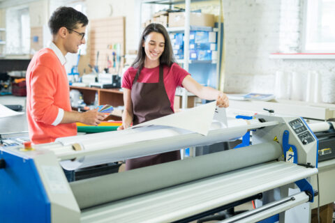 Cheerful excited lady in apron showing printed banner to manager who picking out color on swatch for printing