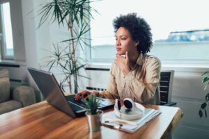 Smiling young African female entrepreneur sitting at a desk in her home office working online with a laptop