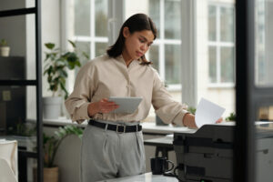 Young confident female office worker in elegant attire printing paper documents on xerox machine and using tablet against window