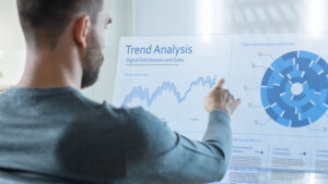 In the Near Future Trade Worker Analyzes Trade Markets on His Transparent Computer Display. Screen Show Interactive User Interface with Statistics, Graphs and Charts. Office is Bright and Modern.