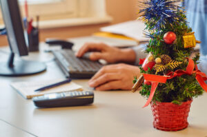 Closeup of desk in office showing small Christmas tree and office worker at computer in background