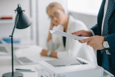Man in foreground holding printed pages from a printer while a woman in the background looks on