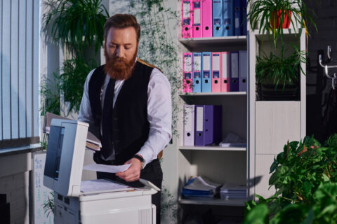 Focused bearded businessman photocopying documents on copier machine at night in office.