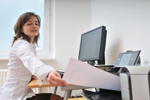 Woman working at desk removing newly printed page from printer alongside