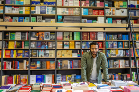 Man in jacket standing in library or book shop with books on shelves with colorful covers and stacks on table