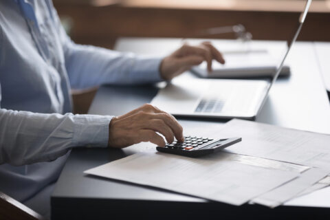 Mature accountant sitting at desk working with stacks of invoices and financial documents, paying utility bills via e-banking on laptop, closeup.