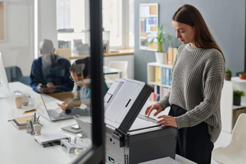 Young woman working in office scanning document, her colleagues using smartphones in background